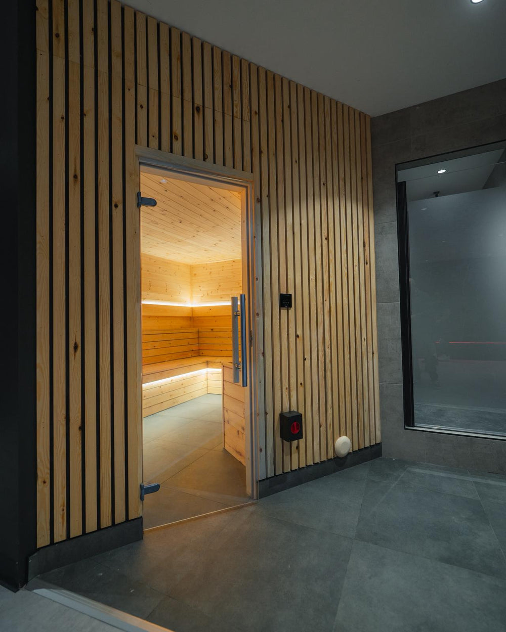 Timber-fronted sauna with a glass door opening onto a tiled changing area